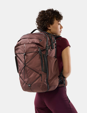 A woman wearing a large garnet hiking backpack, shown from the back and side against a clean light gray studio background.