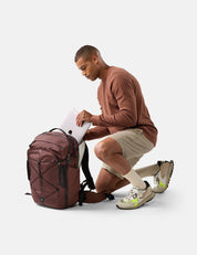 Man putting laptop in garnet backpack, wearing a brown sweater and beige shorts, on a white background