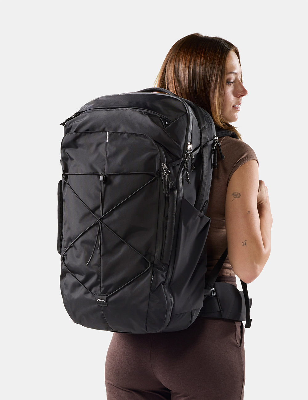 A woman shown from the back wearing a large black backpack against a light gray studio background.