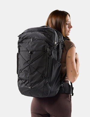 A woman shown from the back wearing a large black backpack against a light gray studio background.