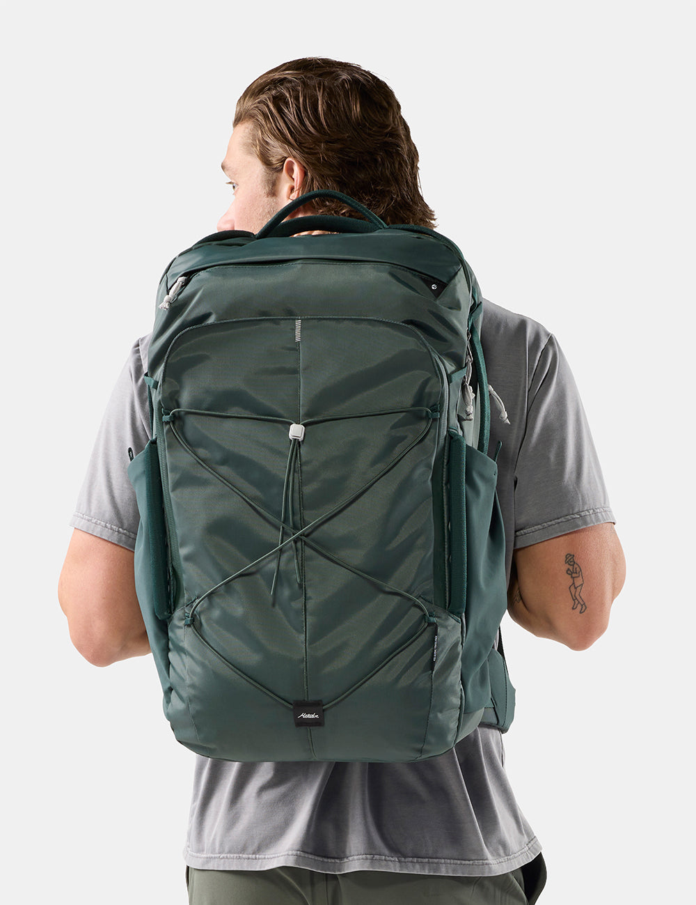 A man viewed from behind wearing a dark green technical backpack with multiple zippered compartments and compression straps, set against a plain light gray studio background.
