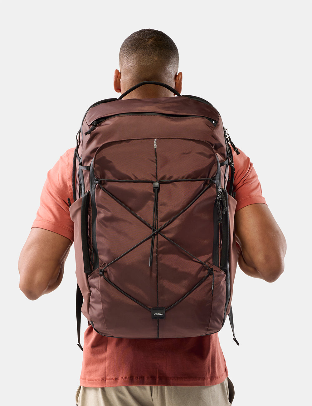 A man viewed from behind wearing a large garnet technical backpack with black compression straps and zippers, over a coral t-shirt, against a light gray background.
