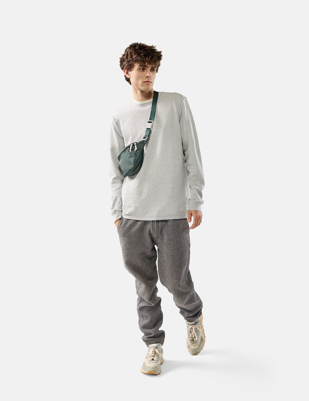 Person wearing a light gray sweatshirt and dark gray pants with a green crossbody bag on a white background