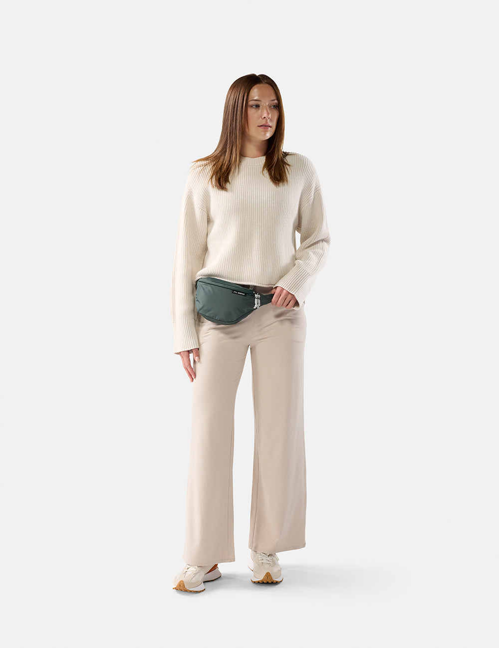 Woman wearing a cream sweater and beige pants with a green fanny pack on a white background