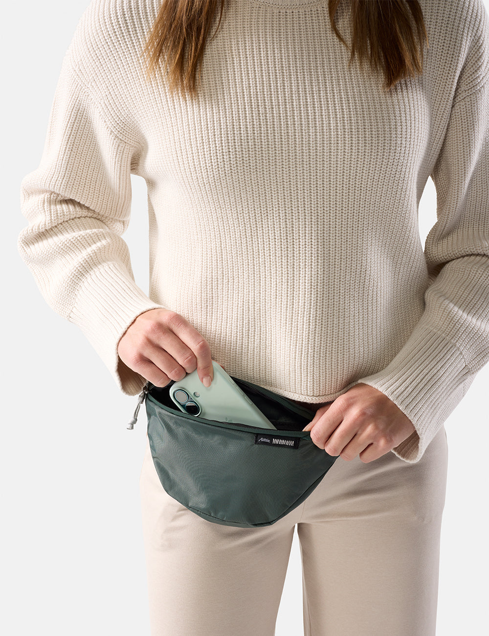 Person wearing a beige sweater and white pants, holding a teal waist bag with iphone