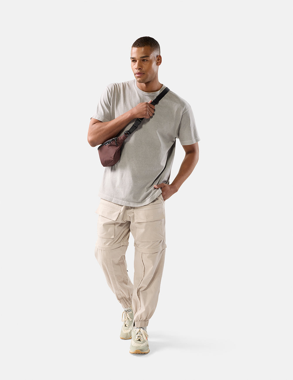 Man wearing a light gray t-shirt, beige pants, and white sneakers, holding a garnet sling on a white background