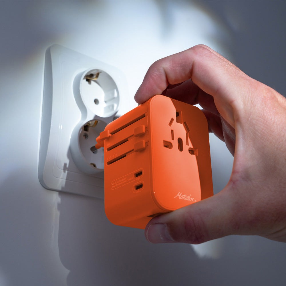 hand holding orange travel adapter, plugging it into an EU outlet on white wall