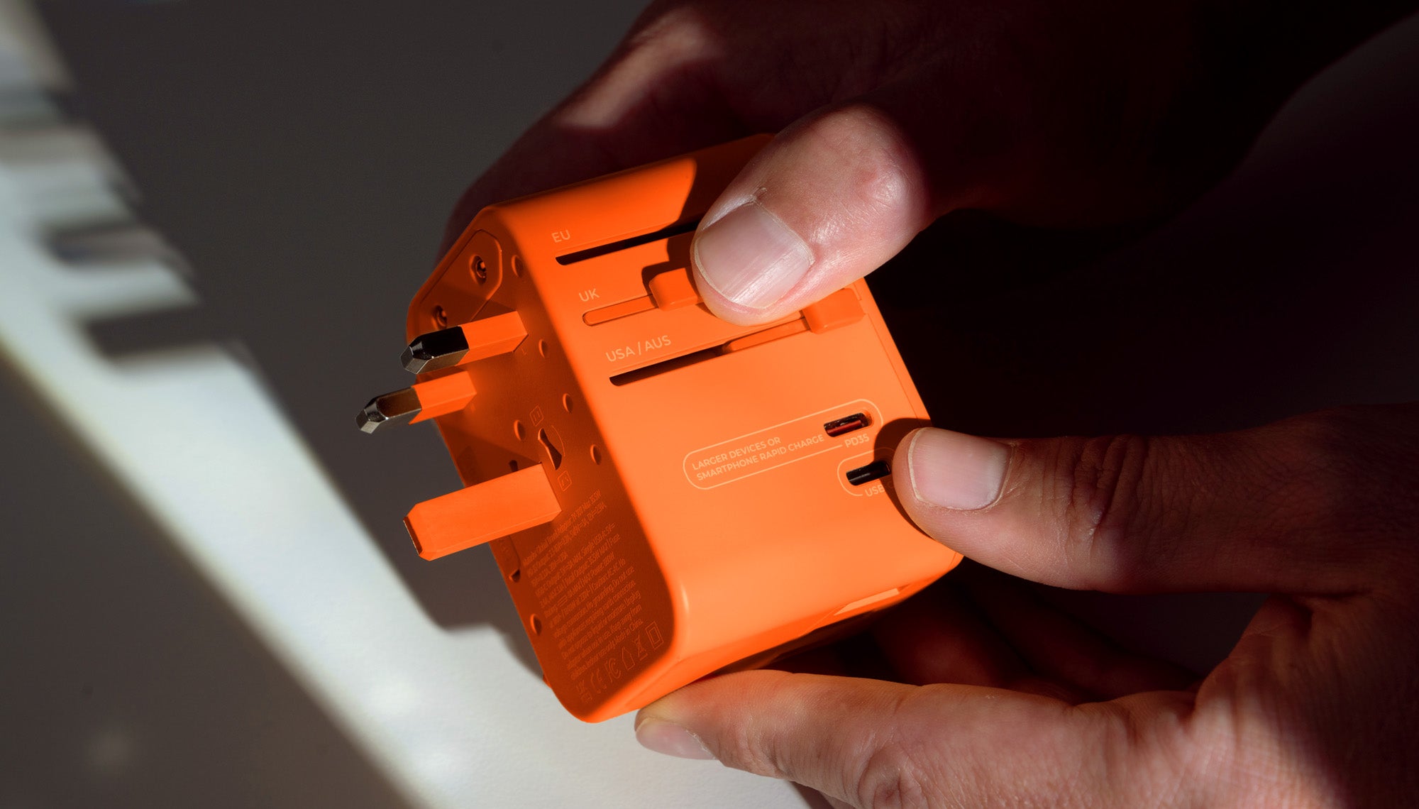 Close up view of orange Travel Adapter. Hand is pushing the UK slider to reveal UK outlet prongs