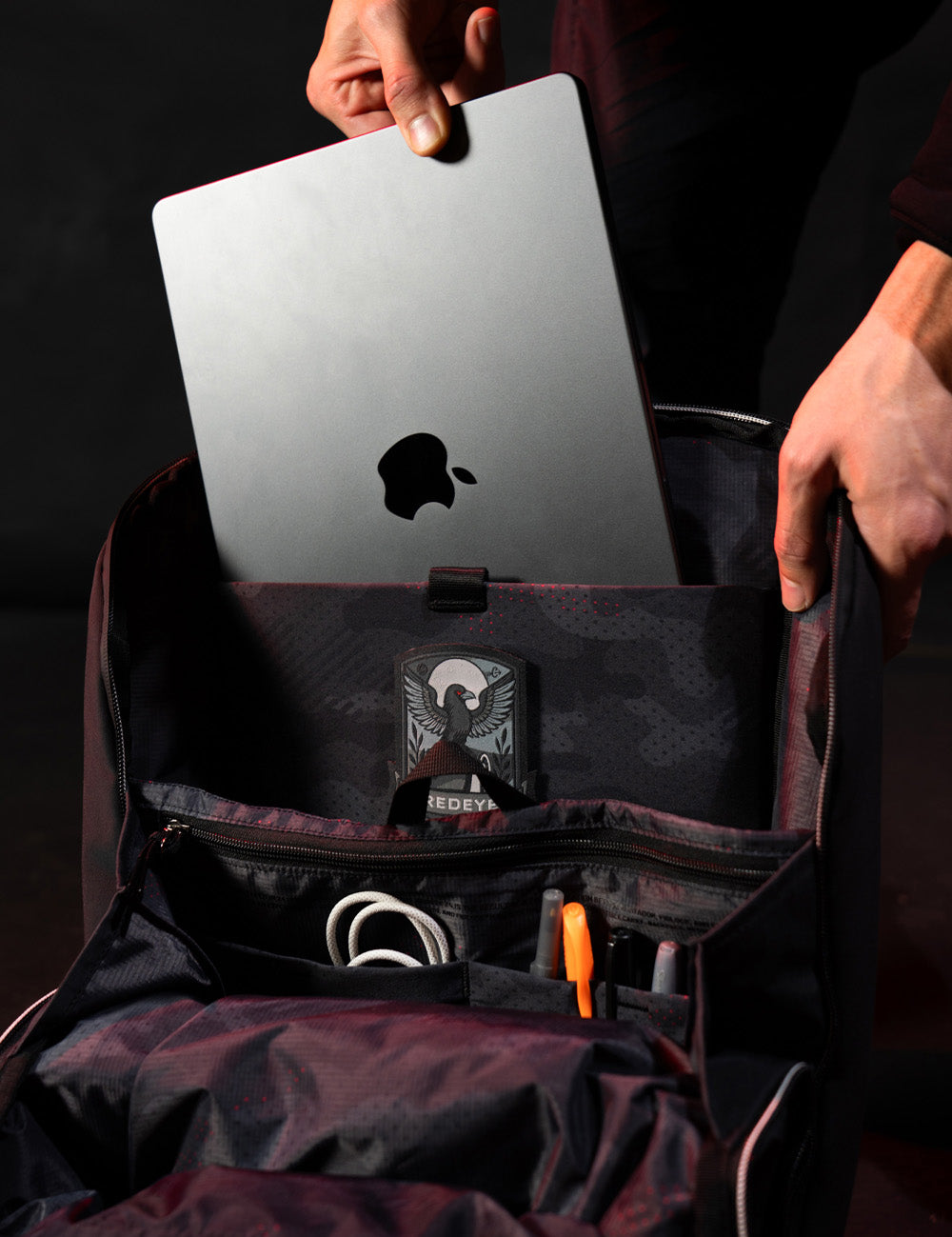 Person placing a silver laptop into a black backpack with visible brand logos.