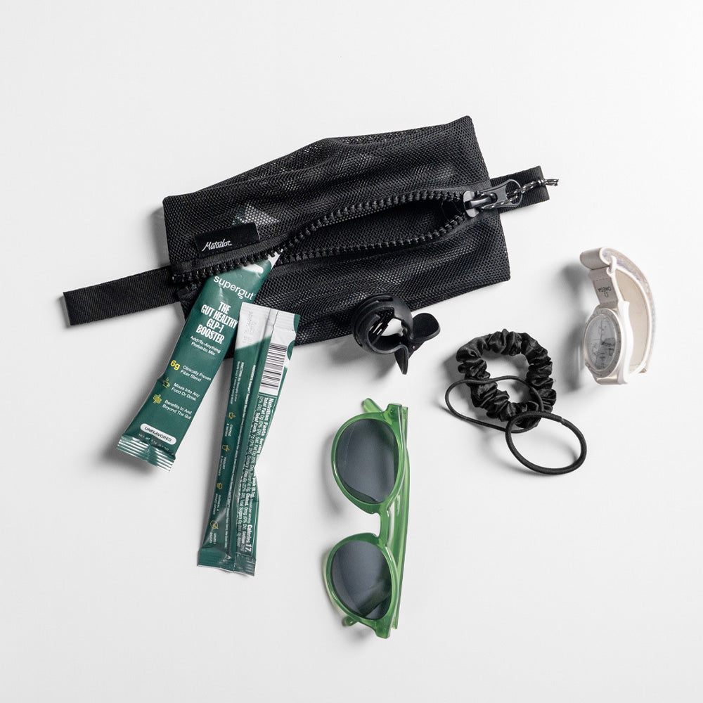 Medium mesh bag on light gray studio background with items spilling out: probiotic packets, sunglasses, watch, and hair ties