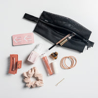 Large mesh bag on light gray studio background with items spilling out: hair ties, hair clips, bobby pins, lip tints, perfume, and contact replacements