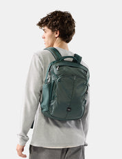 A man wearing a gray sweatshirt and gray pants is shown from the back, wearing a dark green backpack. The photo is taken against a clean, light gray studio background.
