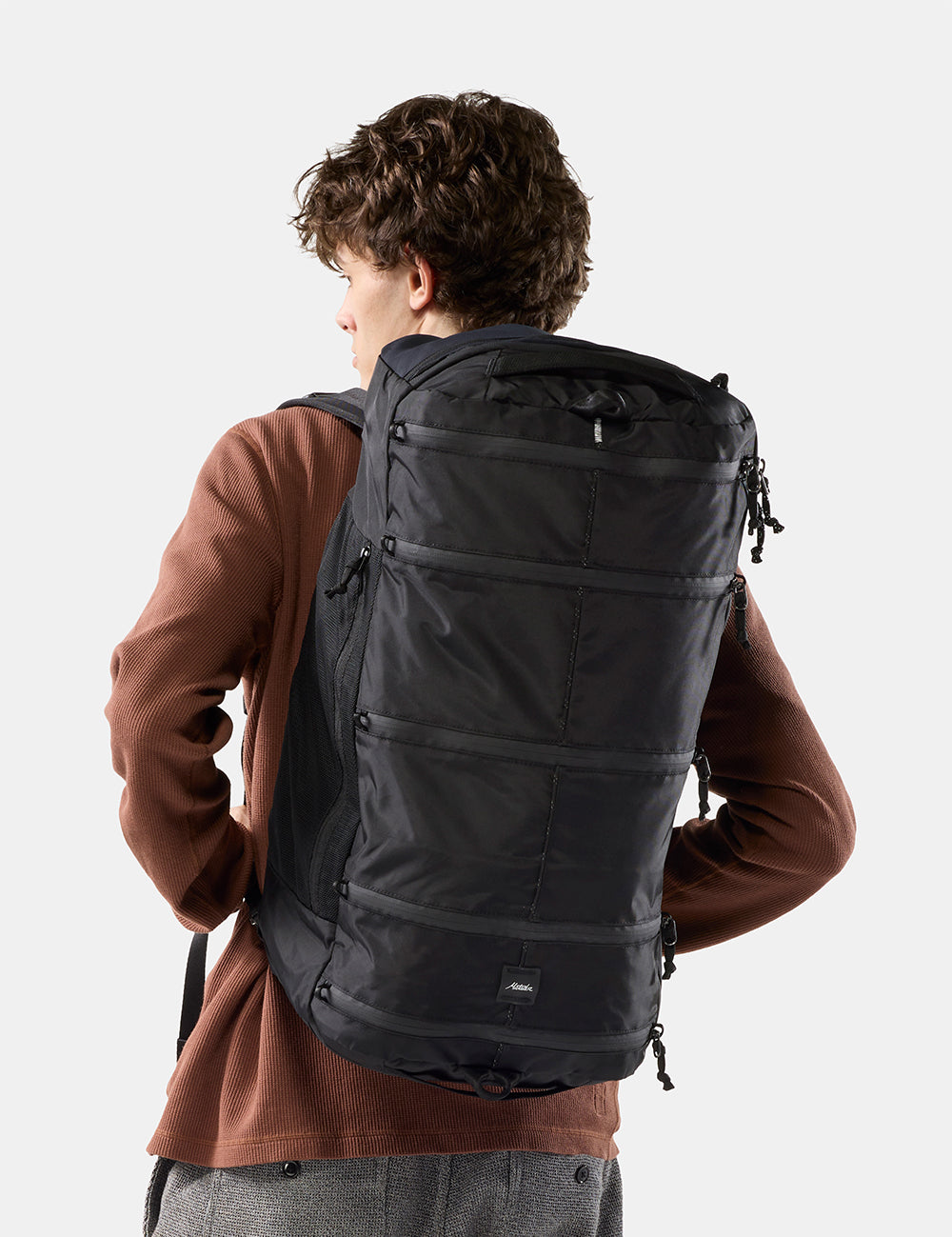 A man wearing a large black backpack, photographed from behind against a light gray studio background.