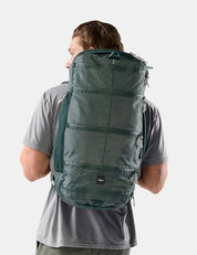 A man wearing a gray t-shirt is shown from behind, carrying a large, dark green technical backpack against a light gray studio background.