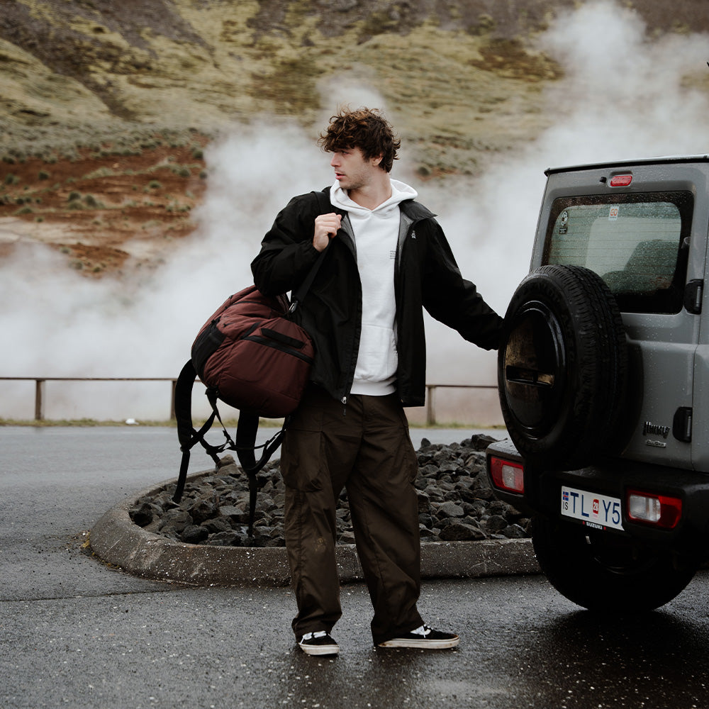 person in moody landscape, holding garnet duffel next to overland vehicle