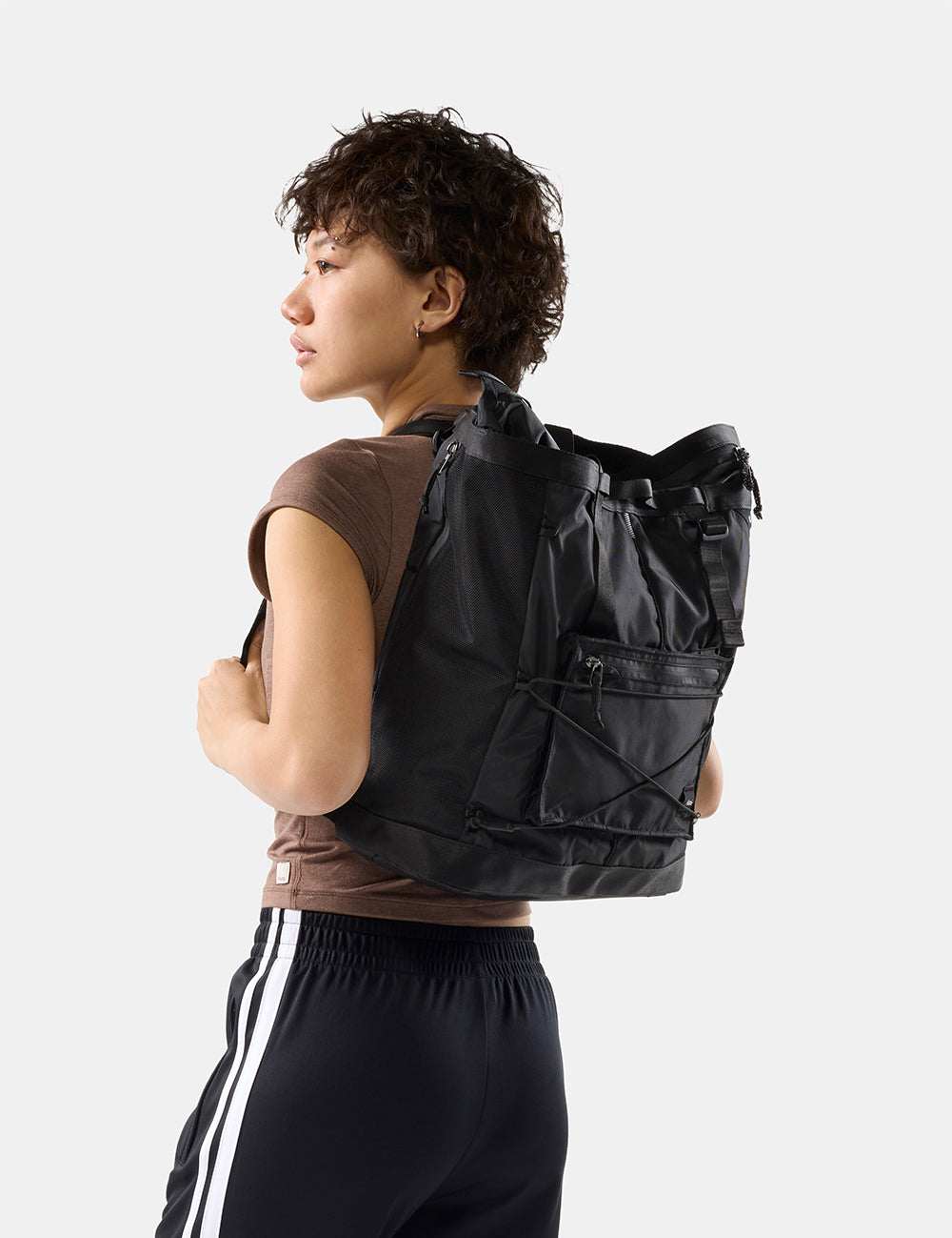 A person wearing a black backpack with multiple zippered pockets and compression straps. They are wearing a brown sleeveless top and black athletic pants with white stripes, photographed against a clean light gray studio background.