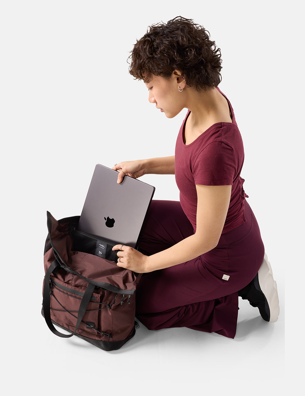 Woman in a burgundy dress placing a laptop into a garnet backpack on a white background