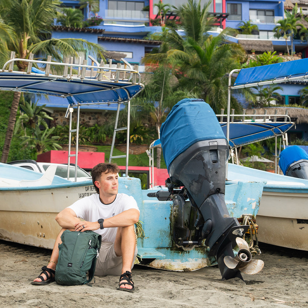 person sitting next to boat on beach, holding juniper backpack