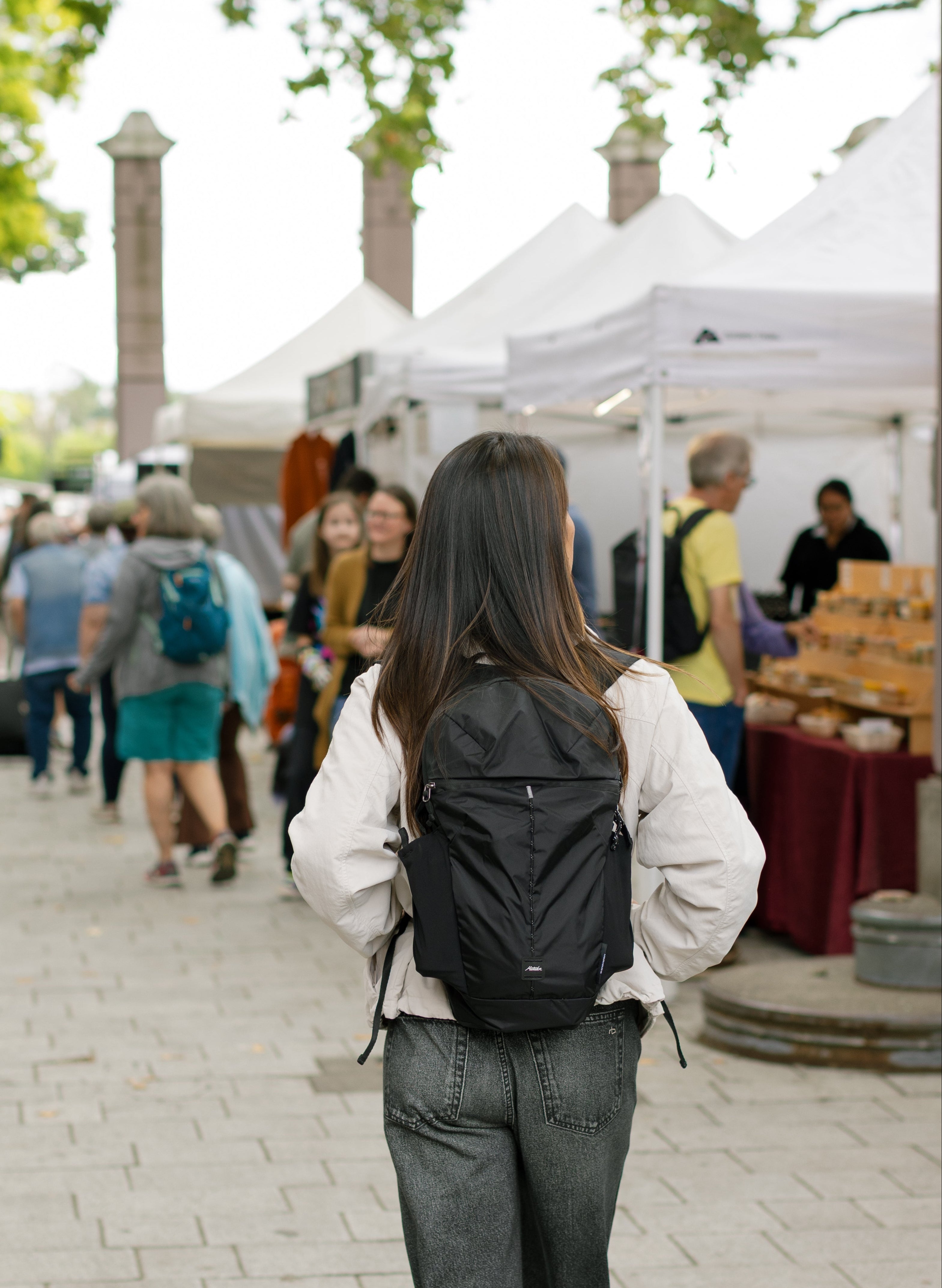 A woman seen from behind walking on a paved outdoor market path, wearing a white long-sleeve top, wide-leg jeans, and a black backpack.