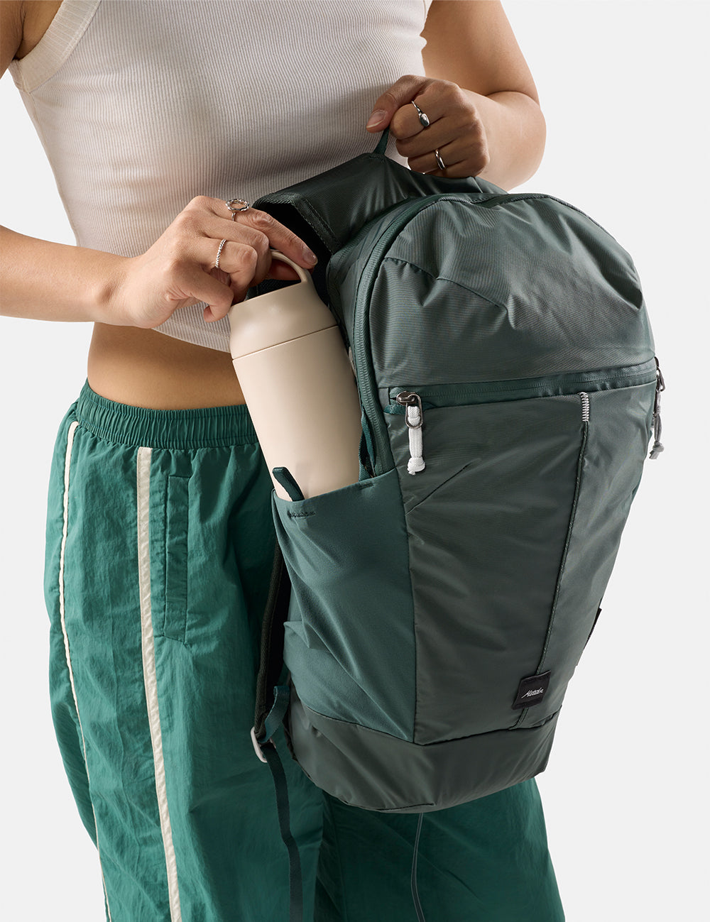 Person holding a green backpack with a white water bottle, wearing a white top and green pants.