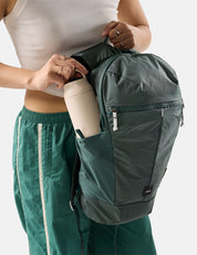 Person holding a green backpack with a white water bottle, wearing a white top and green pants.