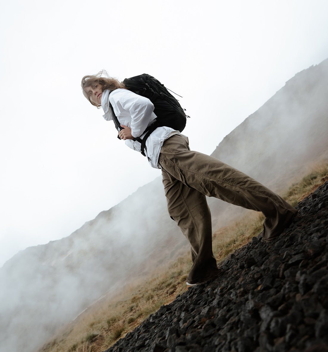 A person hiking on a rocky mountain slope wearing a white shirt, brown pants, and a black backpack, with misty mountains in the background.