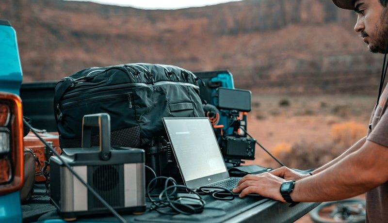 Man in desert setting working on laptop from back of his truck, filled with bags and equipment
