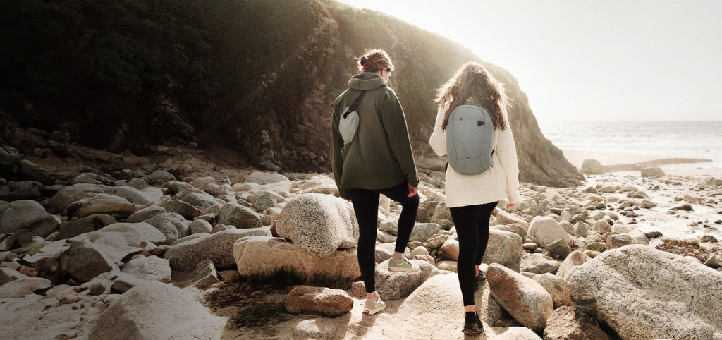 Two women on rocky shoreline, wearing refraction backpack and sling