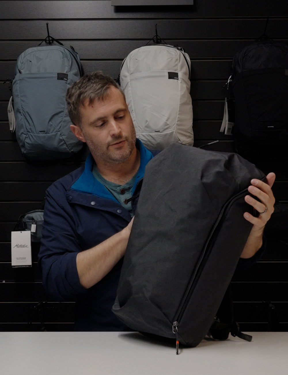 Man standing in front of wall of backpacks holding a black backpack