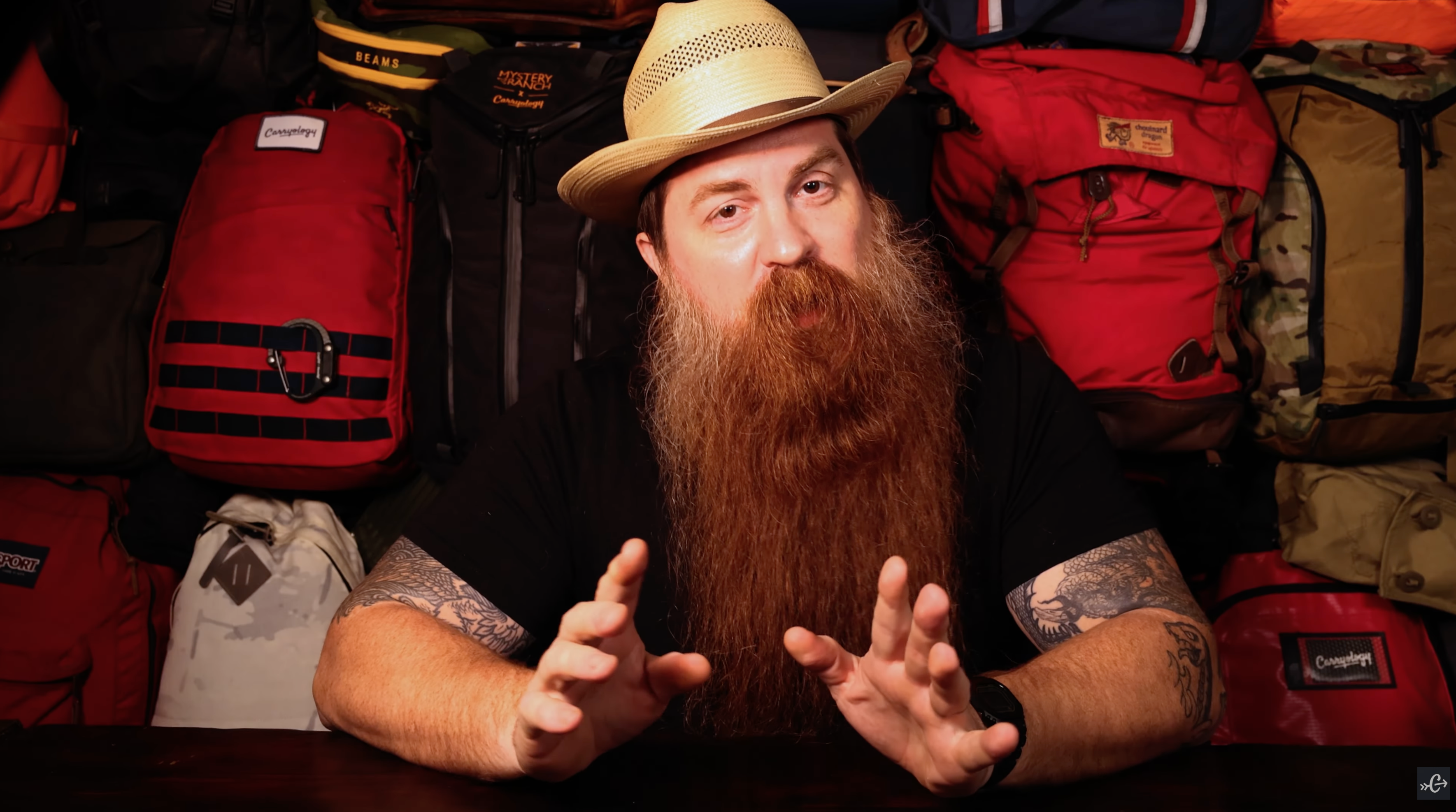 Bearded man in hat sitting in front of wall of backpacks