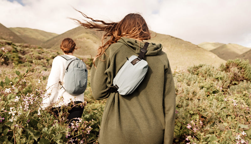 two women, wearing backpack and crossbody sling, walking through a rolling field of wildflowers
