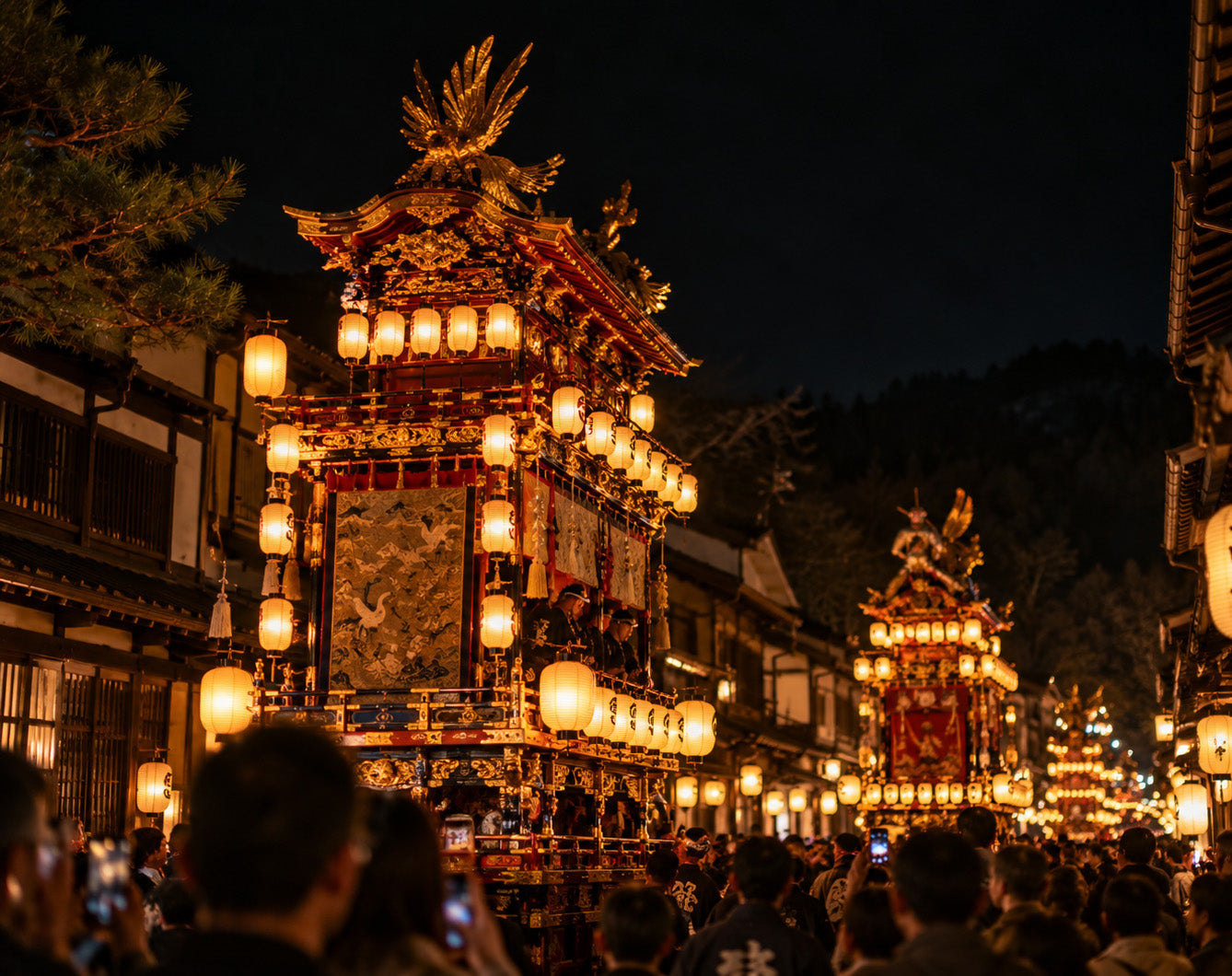 A nighttime street scene showing ornate Japanese festival floats illuminated by many warm yellow lanterns, with a crowd of people in the foreground and dark buildings and hills in the background.