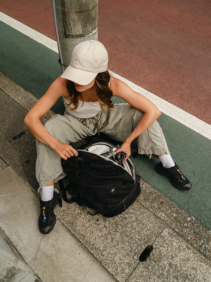A woman sits on a concrete curb next to a basketball court, opening a black backpack. She is wearing a beige baseball cap, a white tank top, light olive cargo pants, white socks, and black sneakers.