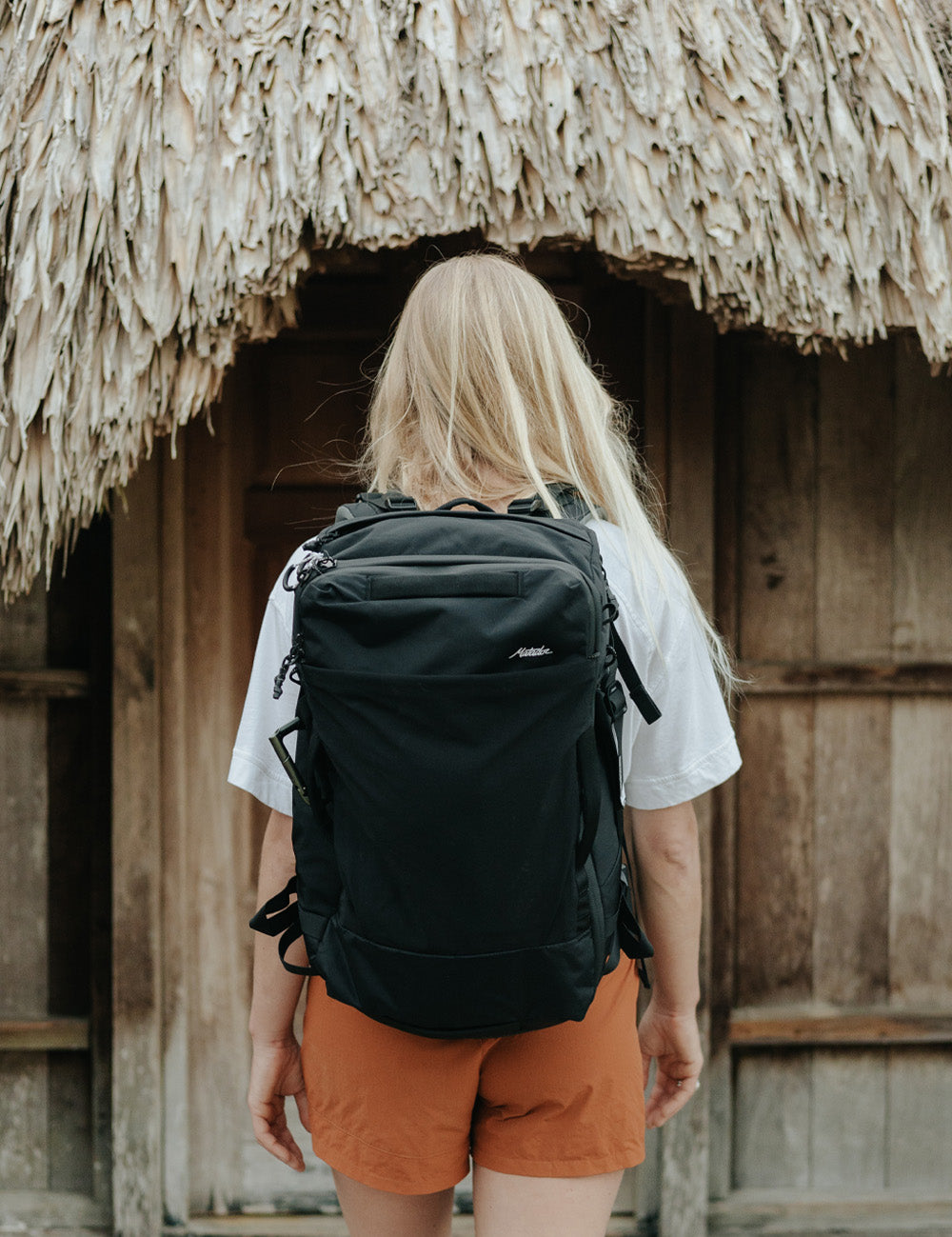 Woman wearing black backpack, walking into grass roof hut