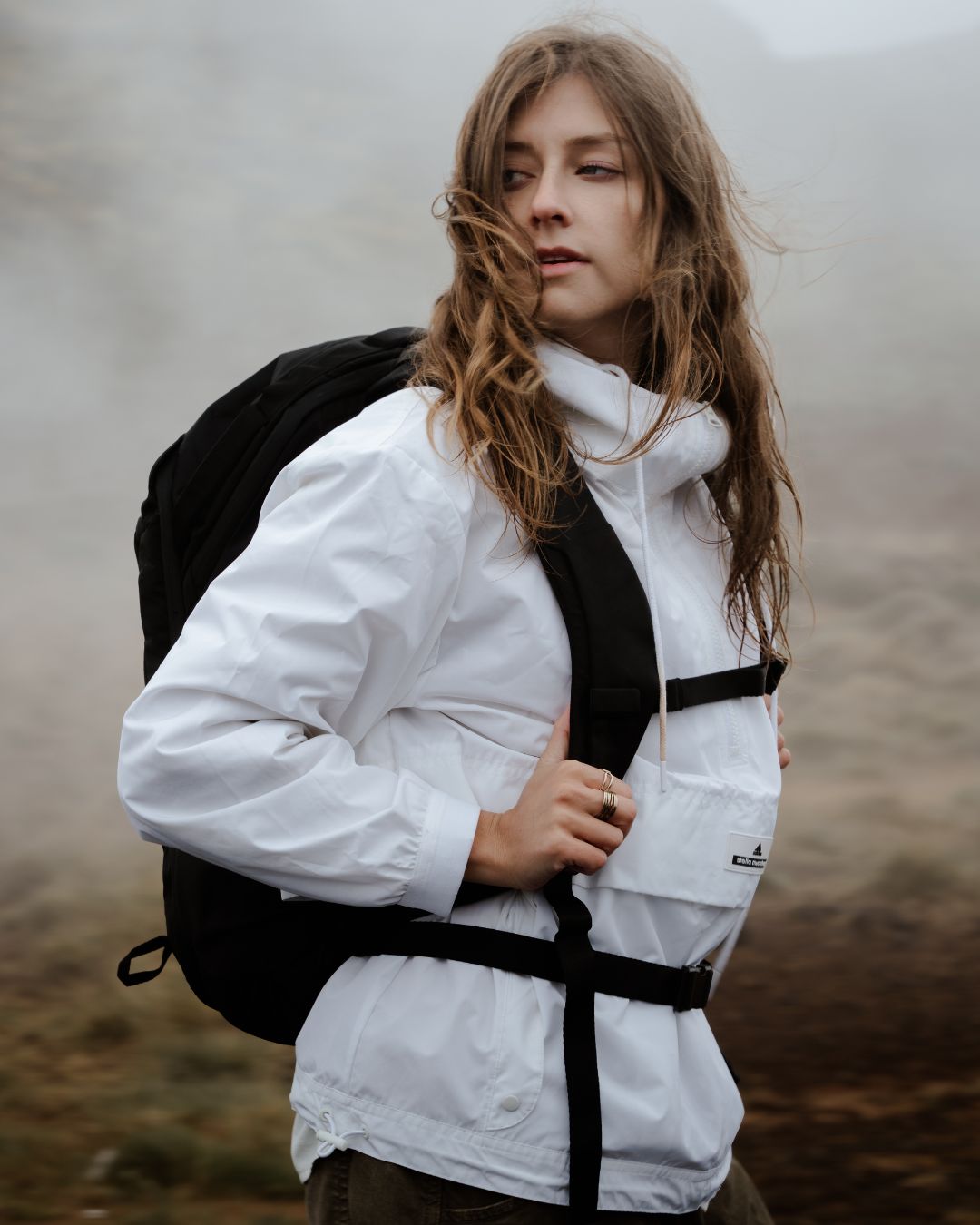 A woman wearing a white hooded technical jacket and a black backpack, standing outdoors in a misty, blurred natural landscape.