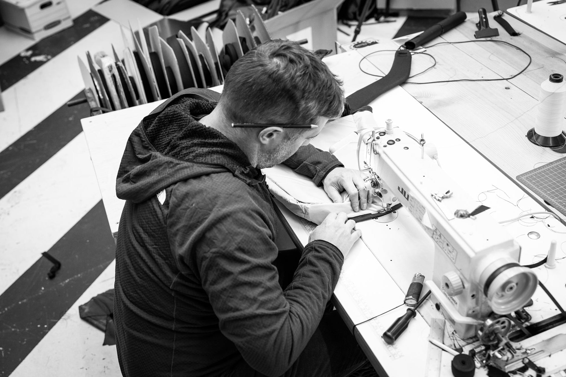 A black and white photograph of a man wearing a dark hoodie, working at a sewing machine in a workshop environment.