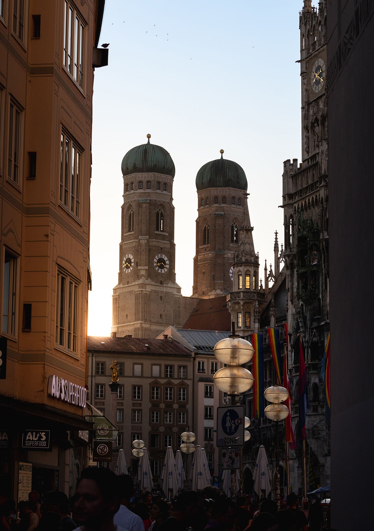 A narrow city street framed by tall buildings, looking toward the twin domes of a historic cathedral at sunset. Street lamps glow, flags hang on the right, and a crowd is visible along the bottom edge.