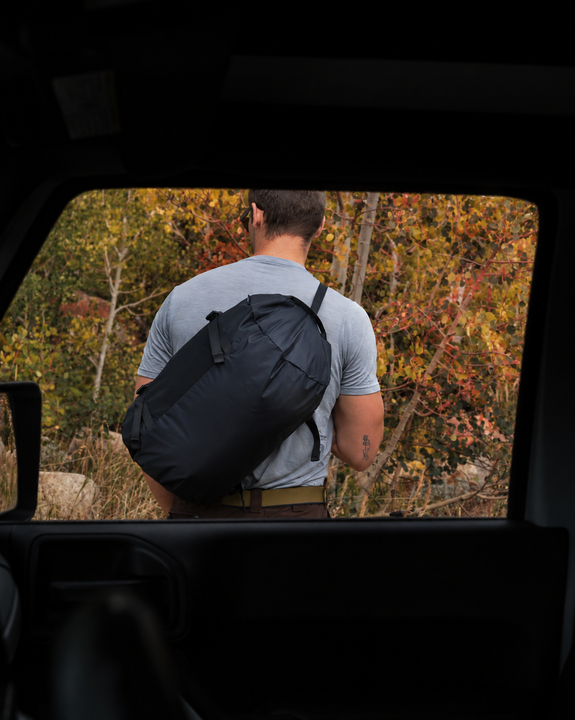 A man viewed from behind wearing a light gray t-shirt and carrying a black duffel bag while standing outside a vehicle with autumn trees in the background.