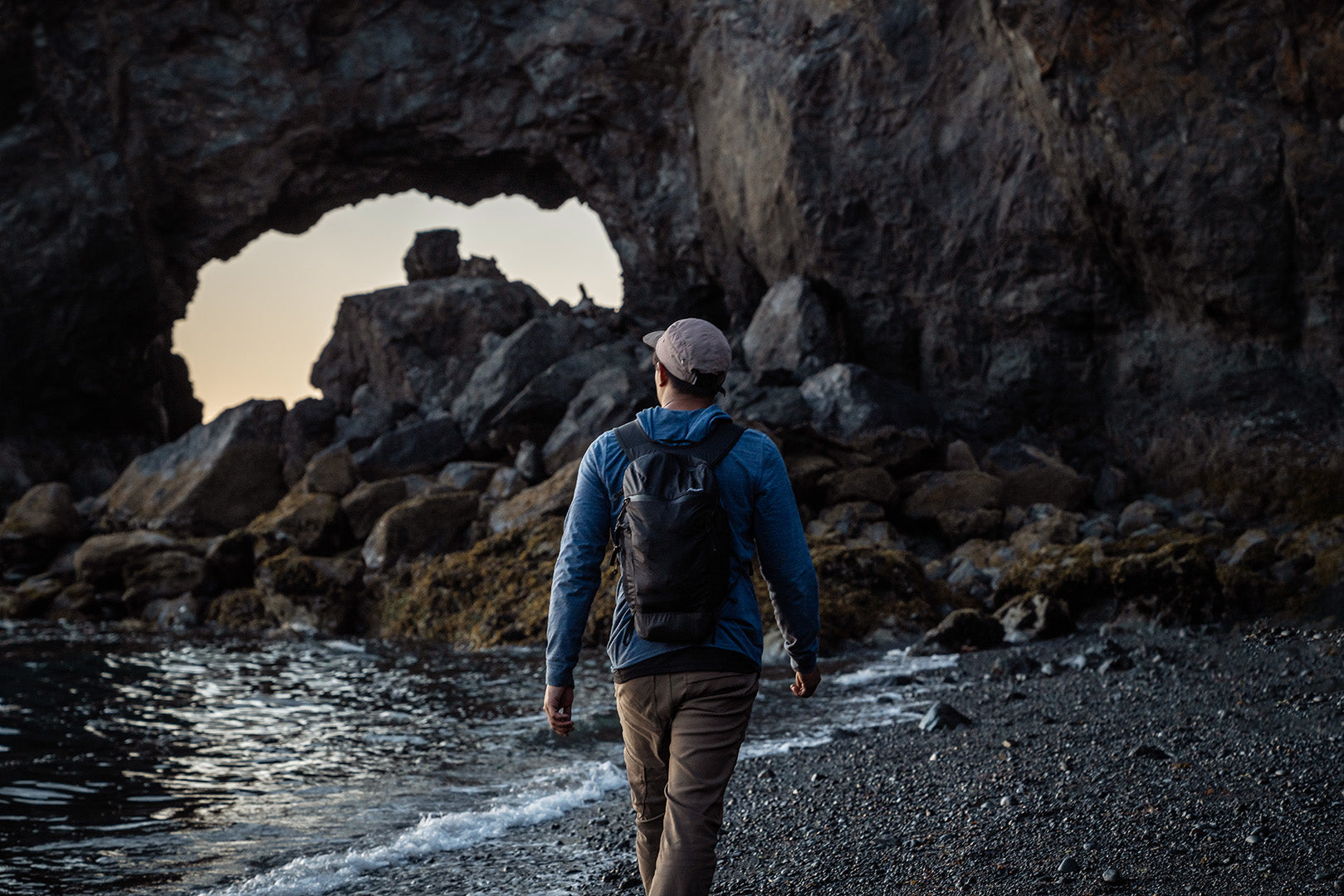 a person wearing a weatherproof backpack walking on a black sand beach by the ocean
