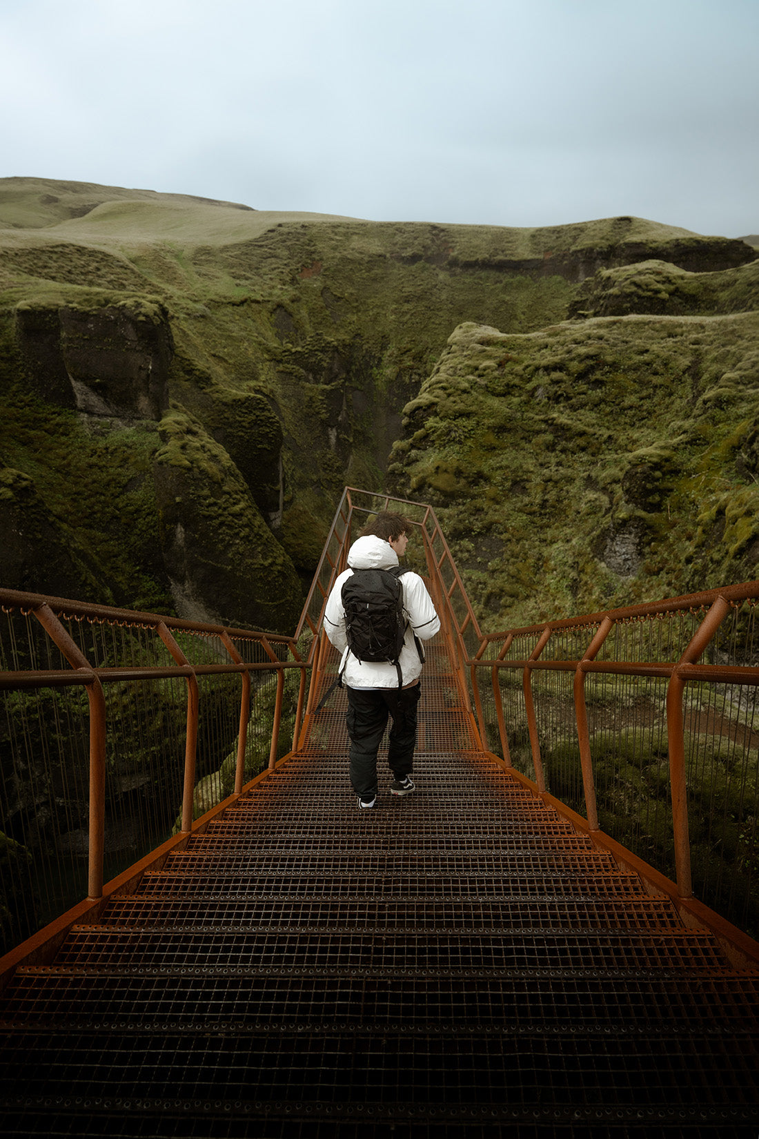 A person with a black backpack walks across a metal staircase in a dramatic outdoor canyon landscape with mossy green cliffs under an overcast sky.