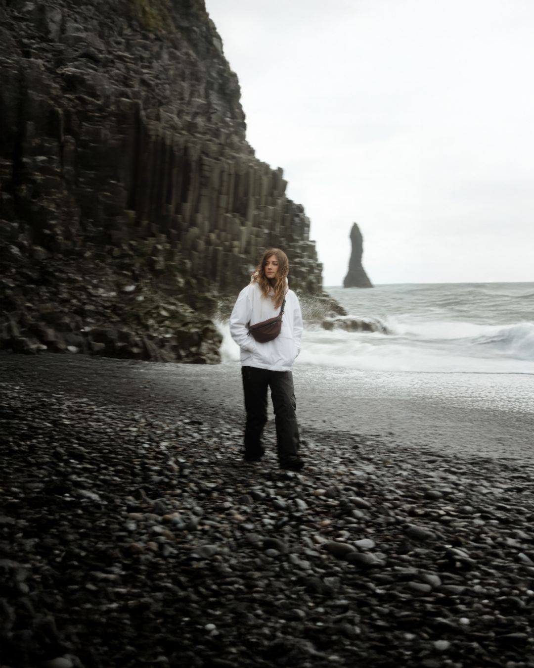 A person standing on a black sand beach in Iceland with a tall basalt cliff on one side and the ocean with a sea stack in the distance, wearing a dark red crossbody bag.