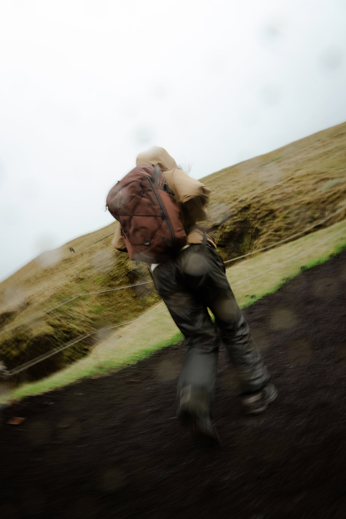 A person hiking up a grassy slope wearing a dark red backpack, dark pants, and a beige jacket, photographed from behind with a motion-blurred outdoor background.