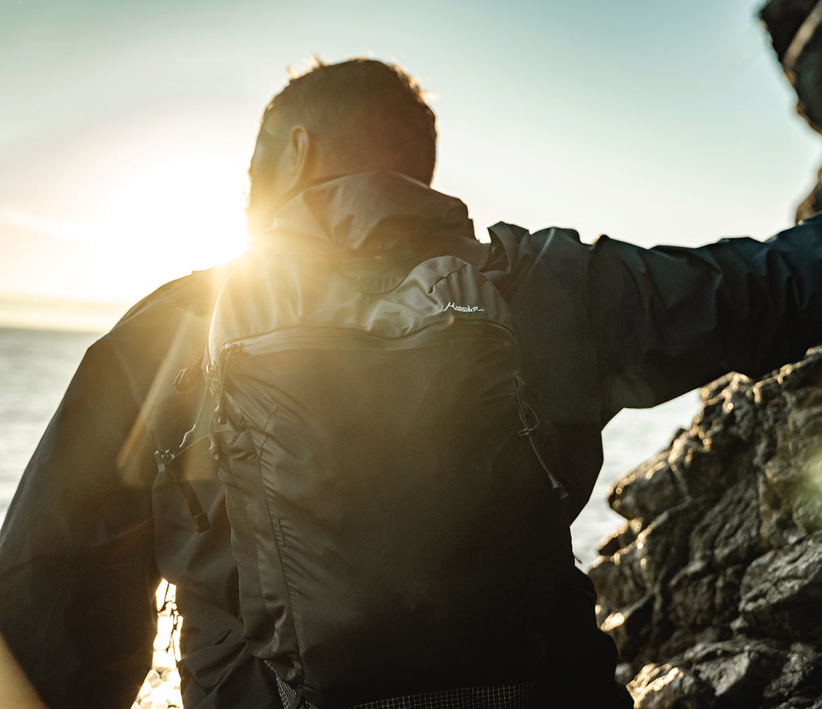 Person wearing an outdoor adventure pack while standing on a rocky outcrop with the sun shining through.