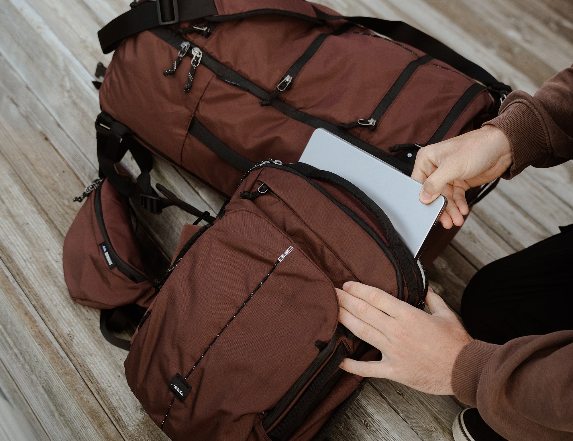 A garnet sling bag and duffel bag with black straps and zippers, shown on a wooden floor with a person's hands placing a silver laptop into a smaller backpack in front.