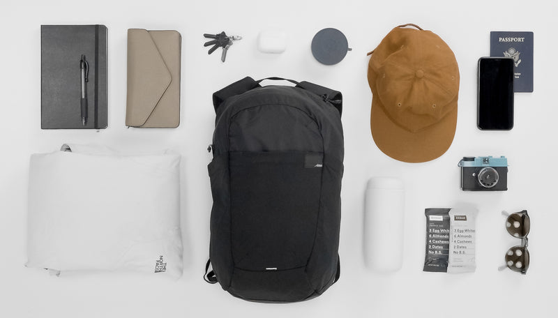 Black backpack on light gray background laid out with various items to go inside: notebook, wallet, rain coat, keys, headphones, canister, hat, waterbottle, RX bars, camera, sunglasses, phone, and passport