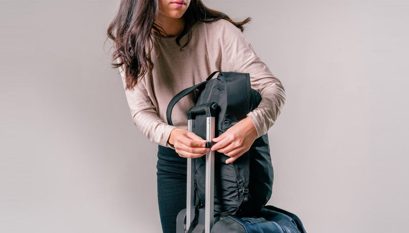 woman on light gray background attaching black backpack to luggage handle
