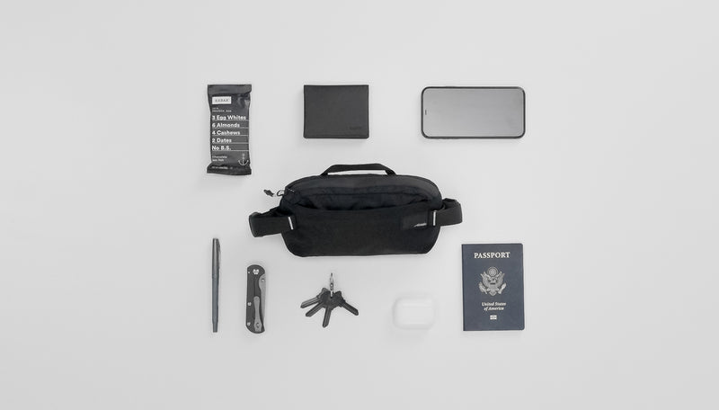 Black sling on light gray background laid out with various items to go inside: RX bar, wallet, phone, pen, pocket knife, keys, headphones, and passport