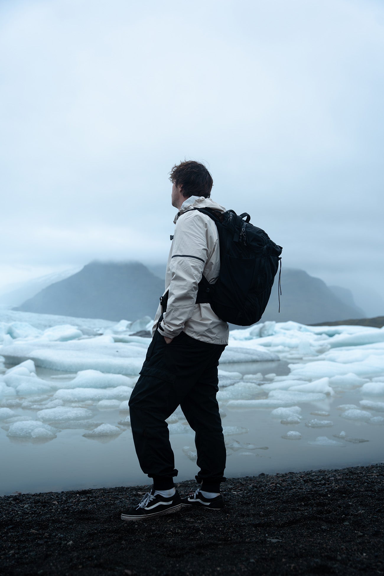 A man wearing a light jacket, black pants, and a black backpack stands on a dark shore, looking out over a glacier landscape with water and floating ice.