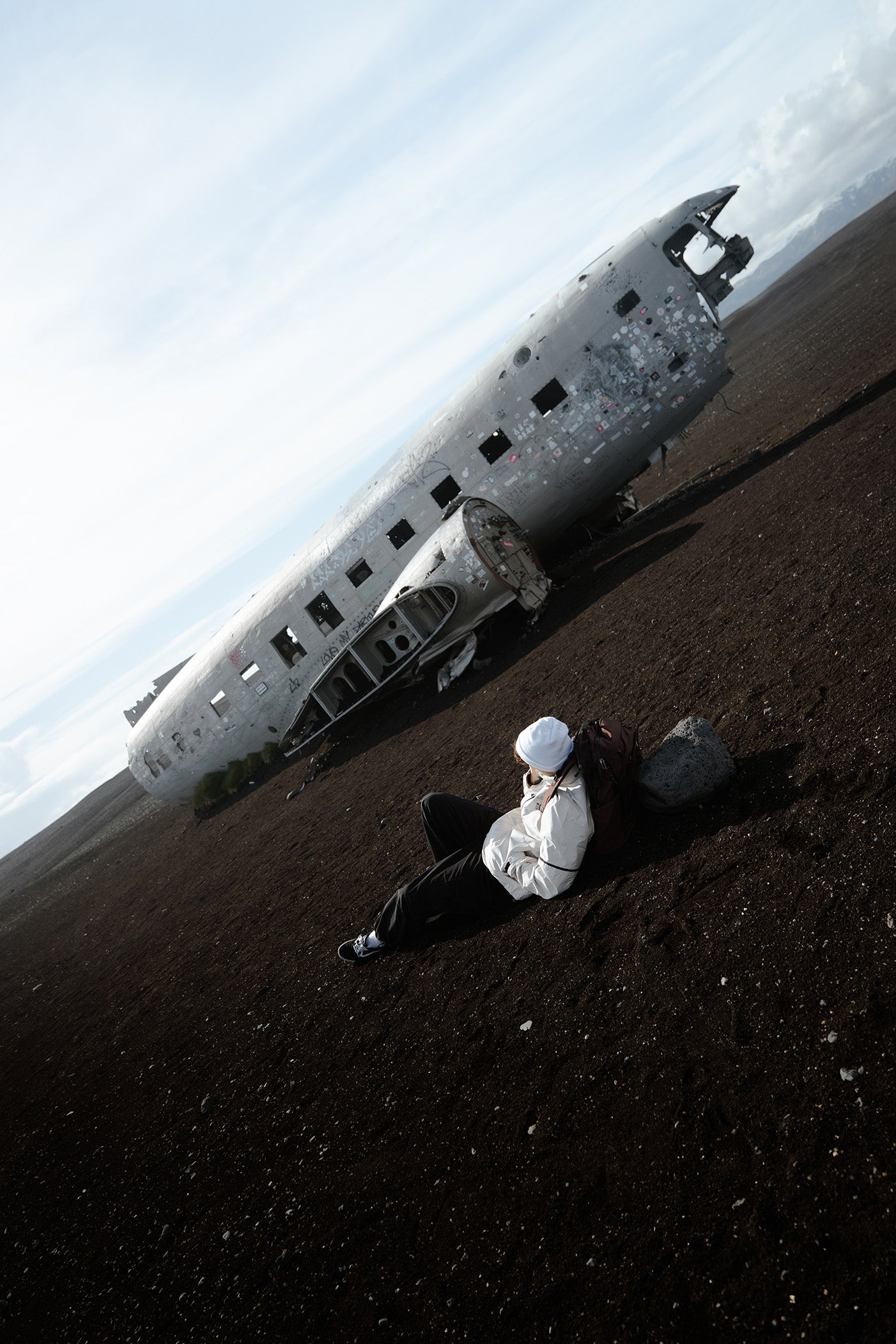 A person in a white rain jacket and black pants lies on the dark volcanic ground in front of the famous airplane wreck site in iceland.