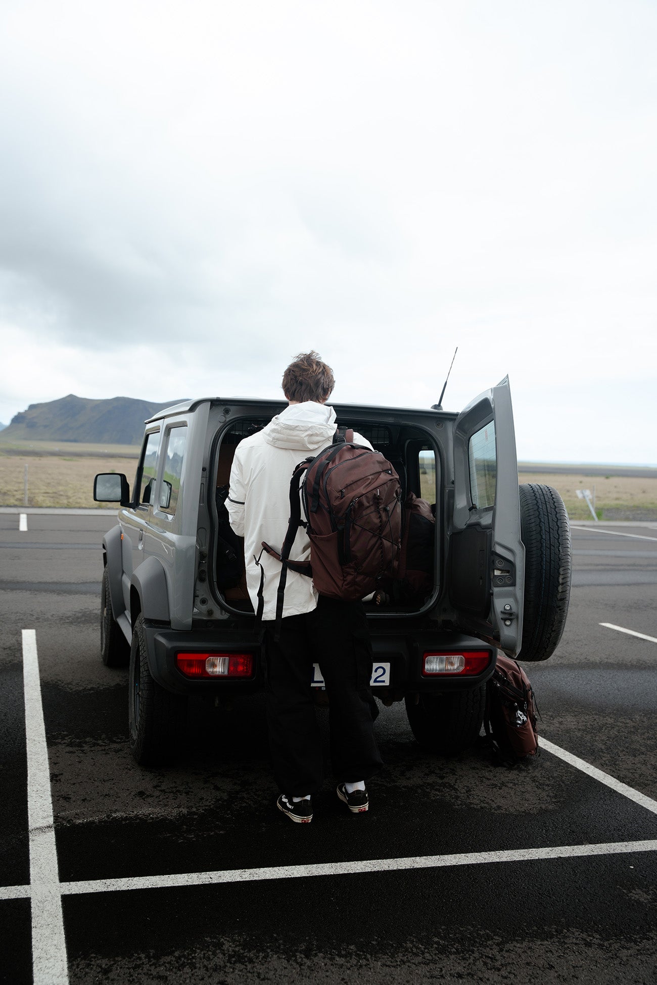 A person in a white hoodie and black pants stands with their back to the camera, loading a garnet backpack into the open trunk of a gray SUV parked in an open paved lot under an overcast sky.